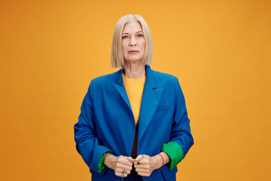 Horizontal medium portrait of stylish senior woman wearing blue jacket standing against bright yellow wall background looking at camera