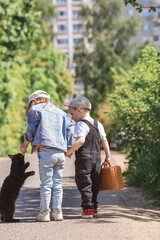 Obraz premium A little girl and a boy in stylish clothes play with a black cat outdoors. The concept of pet care. Children and animals.
