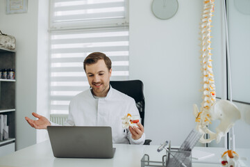 Young male doctor in uniform and eyeglasses showing vertebra model while speaking about spinal...