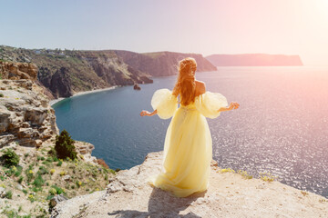 Woman yellow dress sea. Side view Young beautiful sensual woman in yellow long dress posing on a rock high above the sea at sunset. Girl in nature against the blue sky