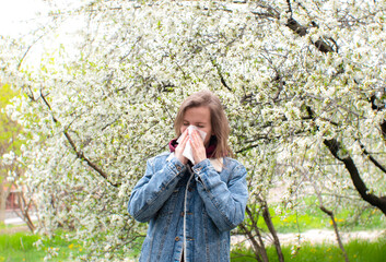 Woman sneezing into a handkerchief near a tree full of blossoms. She is suffering from seasonal...