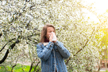 Woman sneezing into a handkerchief near a tree full of blossoms. She is suffering from seasonal...