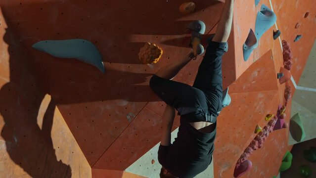 A young millennial man in dark clothes is bouldering on the orange wall of an artificial climbing wall with stones inside. Extreme sports in the evening. High quality 4k footage