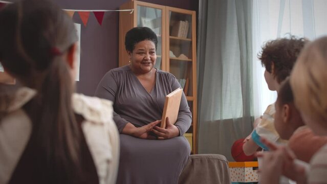 Medium shot of mature African American woman with thick text book in hands discussing topic with multicultural first grade pupils during lesson in bright modern classroom