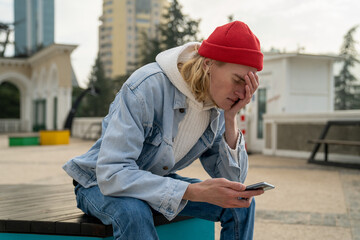 Worried upset millennial guy sitting alone on bench outdoors with smartphone in hands, touching forehead, receiving bad via sms, breaking up with girlfriend over phone, feeling sad and heartbroken