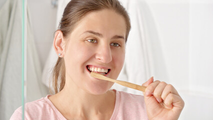 Smiling brunette woman brushing teeth with wooden toothbrush in the bathroom. Concept of teeth health, self checking mouth and oral hygiene.