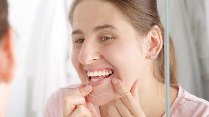 Closeup portrait of young woman checking her teeth at mirror and using dental floss. Concept of teeth health, self checking mouth and oral hygiene.
