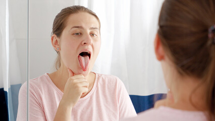 Portrait of young woman checking her teeth and tongue for plaque at mirror in bathroom. Concept of...
