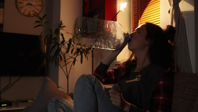 Caucasian tired woman sitting on sofa drinks water from large bottle that is empty. Home evening. Sickness and thirst