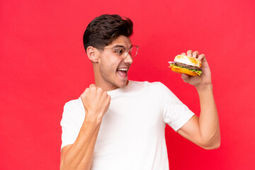 Young Caucasian man holding a burger isolated on red background celebrating a victory