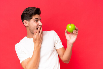 Young caucasian man with an apple isolated on red background with surprise and shocked facial expression