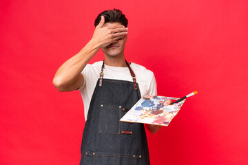 Young artist caucasian man holding a palette isolated on red background covering eyes by hands. Do not want to see something