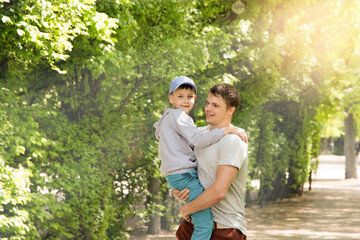 Naklejka na ściany i meble portrait of a cheerful young dad and his son. Dad holds his son in his arms, playing in the park on a sunny summer day. Smile and rejoice