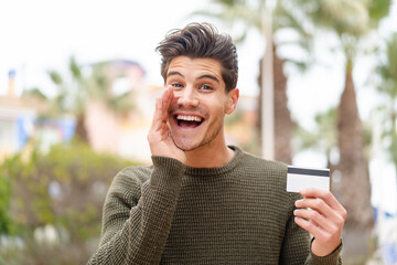 Young caucasian man holding a credit card at outdoors shouting with mouth wide open
