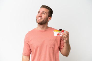 Young caucasian man holding a bowl of fruit isolated on white background looking up while smiling