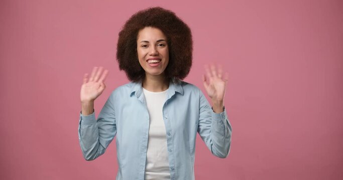 Happy friendly young african american woman waves palms in hello gesture welcomes someone with hospitable expression expresses positive emotions standing over pink background