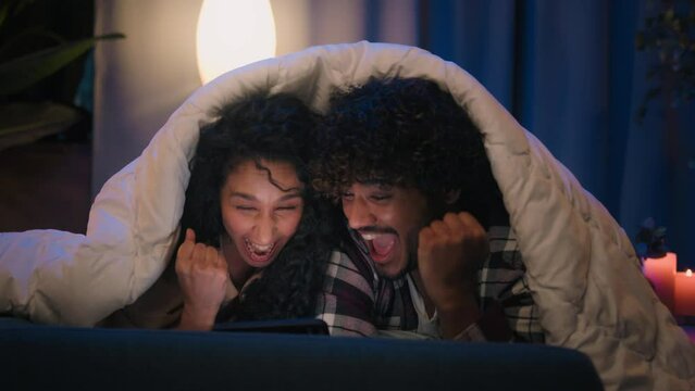 Multiracial couple man and woman at night home win mobile phone bet good news achievement triumph excited with online winning victory smartphone high five at evening couch under blanket gadget addict