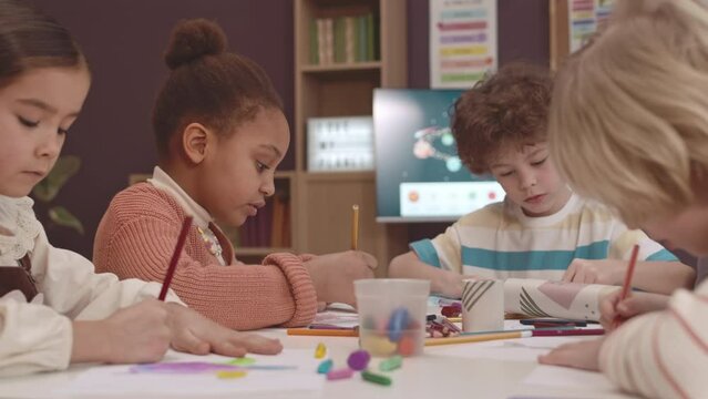 Small group of diverse elementary age kids drawing and crafting together in preschool with teacher helping them