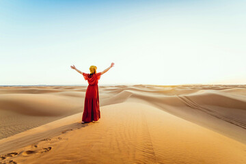 Woman wearing hijab walking in the desert sand dunes at sunset - Happy traveler with arms up enjoying freedom outside - Wanderlust, wellbeing, happiness and travel concept - obrazy, fototapety, plakaty