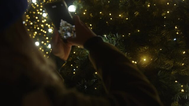 Woman taking pictures of glowing Christmas tree lights with smartphone