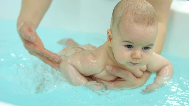 Baby girl splashes hands and kicks legs trying to swim forward in bathtub. Swim teacher pours water on newborn back encouraging swimming reflexes