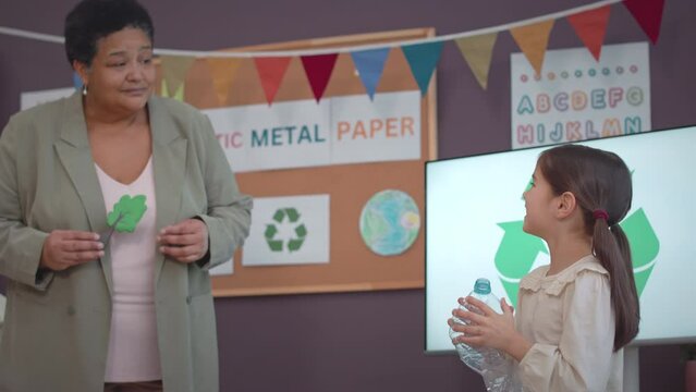 Medium shot of smart elementary age Asian girl with crushed plastic bottle in hands making presentation on recycling and garbage sorting process at school
