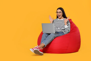 Excited woman with laptop sitting on beanbag chair against orange background