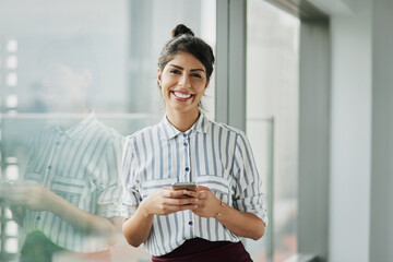 Business woman, portrait and phone with happiness at office window with a smile. Young female face employee and mobile connection of a worker feeling happy on social media and technology at company