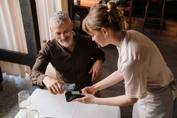 Fototapeta na wymiar Positive young waitress accepts payment through bank terminal for lunch of smiling mature man in cafe