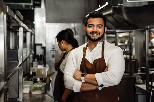 Positive busy indian male business owner in apron looking at camera in cafe kitchen