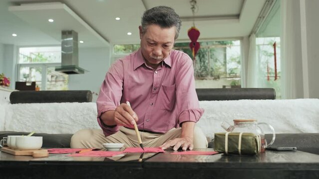 Senior Asian man sitting on sofa in living room and drawing traditional calligraphy on red paper while preparing decor for Tet holiday