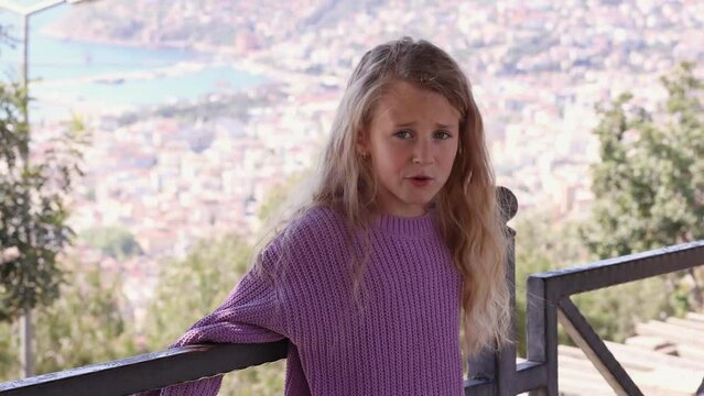 Little girl with long curly blond hair in violet sweater standing outdoors and explaining something looking at camera, true child's emotions
