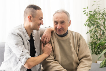 Young adult caucasian son listening and supporting his old elderly senior father at home indoors. Care and love concept.