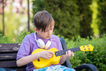  a boy plays a small guitar, a yellow ukulele on a bench in the park