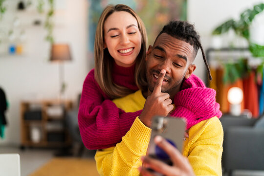 Delighted young multiracial couple in colorful wool sweater smiling and taking selfie on smartphone while sitting at wooden table together in modern apartment