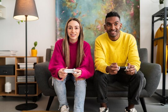 Cheerful multiracial boyfriend and girlfriend in colorful casual clothes sitting on sofa with gamepads and playing video game together in modern living room