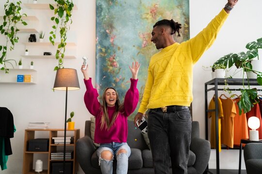 Delighted diverse boyfriend and girlfriend in bright sweaters raising arms and shouting while celebrating success in video game in modern apartment
