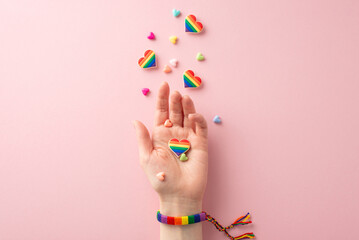 Woman proudly display her support for the LGBT community in this first person top view photo featuring hand adorned with a rainbow bracelet and holding a heart-shaped pins on a pastel pink background