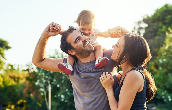 Family, dad and daughter on shoulders in park with mom, happiness or love in summer sunshine. Young couple, baby girl or laugh together for freedom, bond or holding hands for care, backyard or garden
