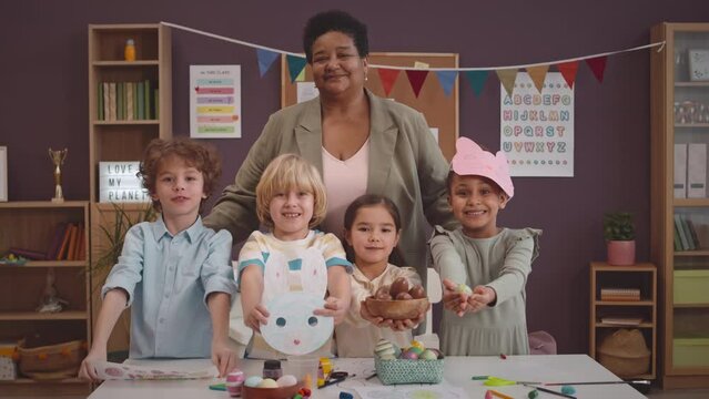 Medium portrait of cheerful mature African American female teacher and group of multicultural primary school kids saying Happy Easter at camera standing in bright decorated classroom