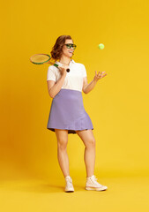 Portrait of young, beautiful girl in sportswear and glasses, smiling posing with tennis racket and ball against yellow studio background. Concept of sport, active lifestyle, emotions, fashion, hobby