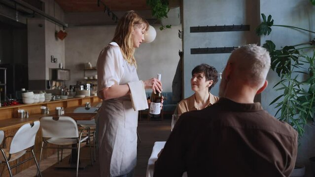 Pretty blonde waitress opens bottle of wine for elderly couple in cafe