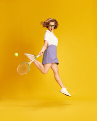 Full-length portrait of young active girl in sportswear jumping with tennis racket against yellow studio background. Concept of sport, active lifestyle, emotions, fashion, hobby