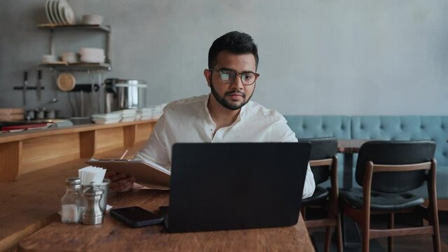 Serious indian male business owner in eyeglasses filling orders on laptop in cafe
