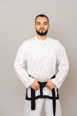 Serious young bearded man wearing taekwondo uniform is holding his black belt.