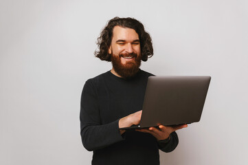 Wide smiling bearded man is holding an opened laptop and typing on it.