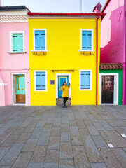 Smiling woman posing in front of yellow house at Burano Island. Happy traveler female tourist with raincoat and sun glasses posing and having fun near colorful houses  in Venetian lagoon.