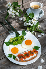 Sandwich with salted trout and avocado for breakfast. Two fried eggs on a plate. A cup of coffee on a wooden table.
