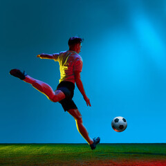 Dynamic portrait of soccer, football player in sports uniform kicking the ball over field, stadium background in neon light. In motion