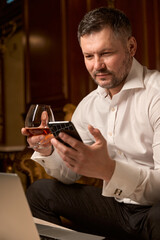 Handsome man with smartphone alone and glass of whiskey in restaurant